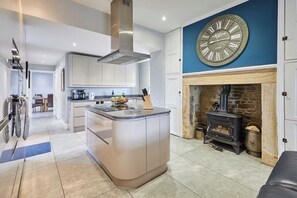 A modern kitchen with a central island, a large wall clock, and a stone fireplace.