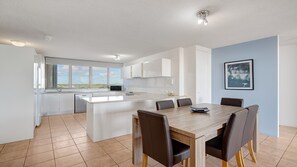 A modern kitchen with white cabinets, a central island, and a dining area with a wooden table and chairs. - Gemini Resort 55 Stunning Ocean Views (Golden Beach)
