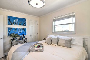 Een slaapkamer met een bed, een raam, kunst aan de muur en een nachtkastje met een vaas en boeken.
