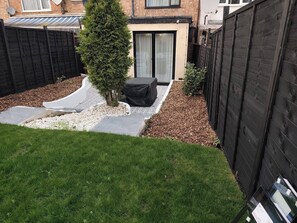 Ein Garten mit Terrasse, einem kleinen Baum und einem schwarzen Zaun.