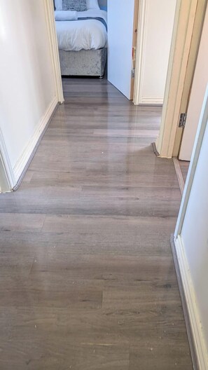A hallway with wooden flooring leading to a bedroom with a bed and a nightstand. - 3 bed House upto 6 Adults (Tilbury)