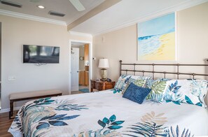 Un dormitorio con una cama grande, televisión, un cuadro de una playa y un baño visible a través de una puerta abierta.