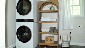 Un espace buanderie comprenant un lave-linge et un sèche-linge à double porte, une étagère en bois avec des serviettes pliées et des paniers à linge, et une fenêtre avec un rideau blanc.