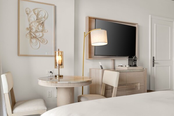 Quarto de hotel com uma mesa redonda, uma cadeira, uma cama, televisão, abajur e uma obra de arte emoldurada na parede.