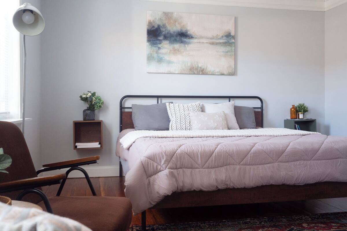 A bedroom with a bed, a chair, a bedside table with a plant, and a painting on the wall.