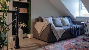 Un dormitorio estilo loft acogedor, con techo inclinado, un sofá gris con cojines estampados, una mesita redonda y una alfombra vibrante con diseño.