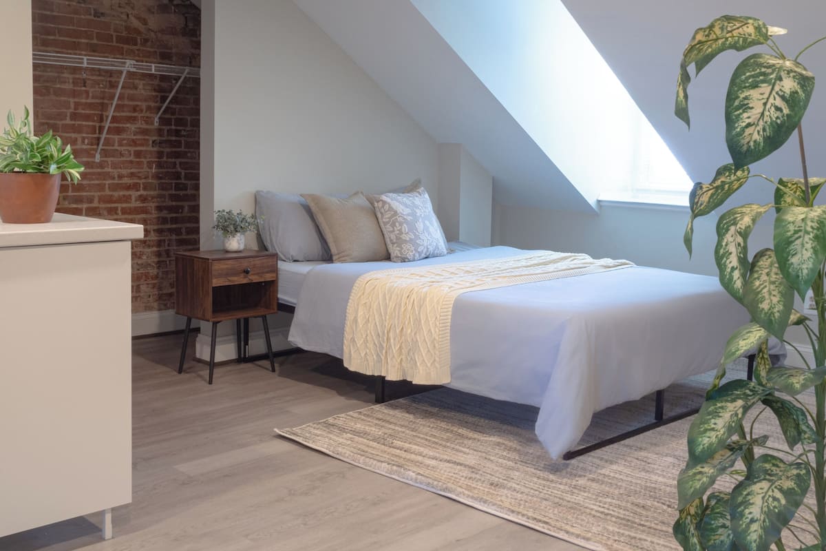 A bedroom with a sloped ceiling, a bed with a white bedspread, a small bedside table with a plant, and a brick wall.