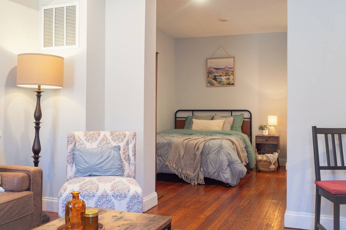 A bedroom with a bed, a chair, a lamp, a bedside table, and a painting on the wall.