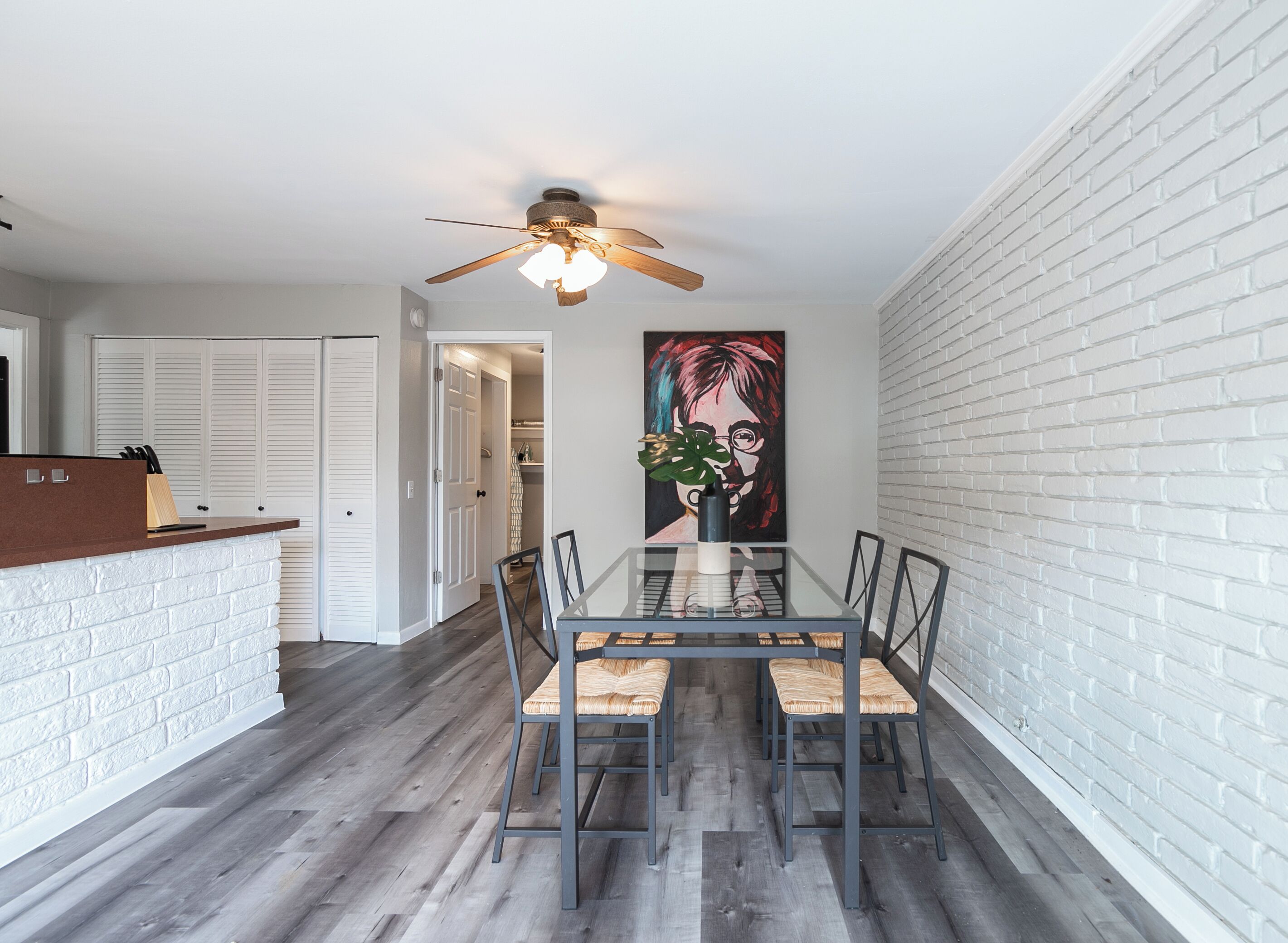 A dining area with a glass table, cushioned chairs, a ceiling fan, and a white brick wall.