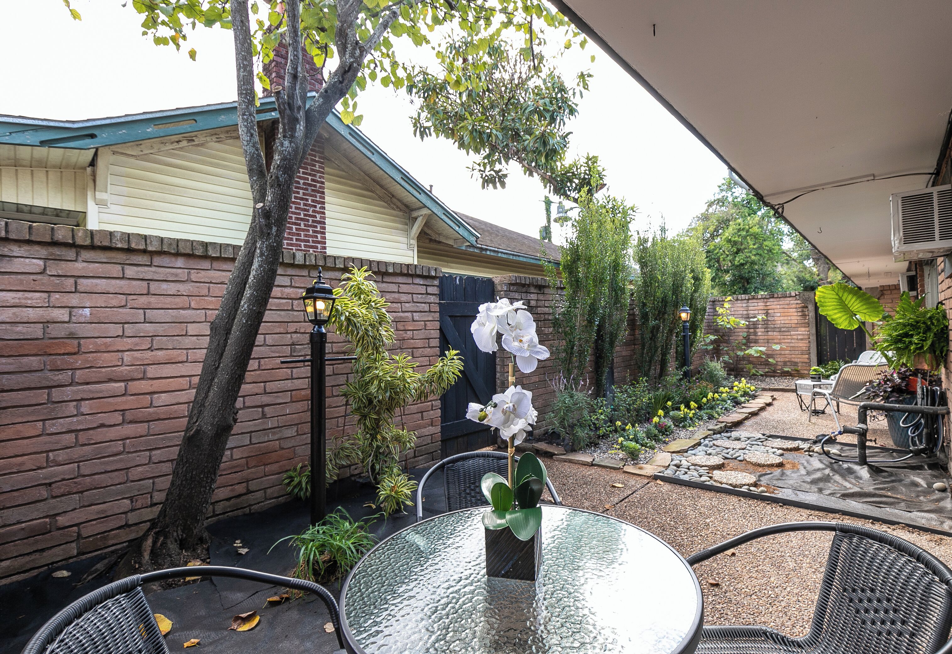 Une table en verre entourée de chaises, placée dans un espace patio à l’arrière de la maison, avec des murs en briques, un arbre et des plantes en pot.