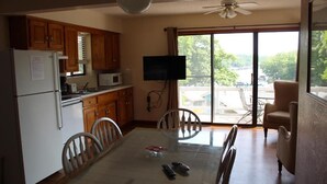 A compact kitchen with wooden cabinets, a refrigerator, a microwave, and a dining table with chairs. A television is mounted on the wall, and there is a sliding glass door leading to an outdoor area.