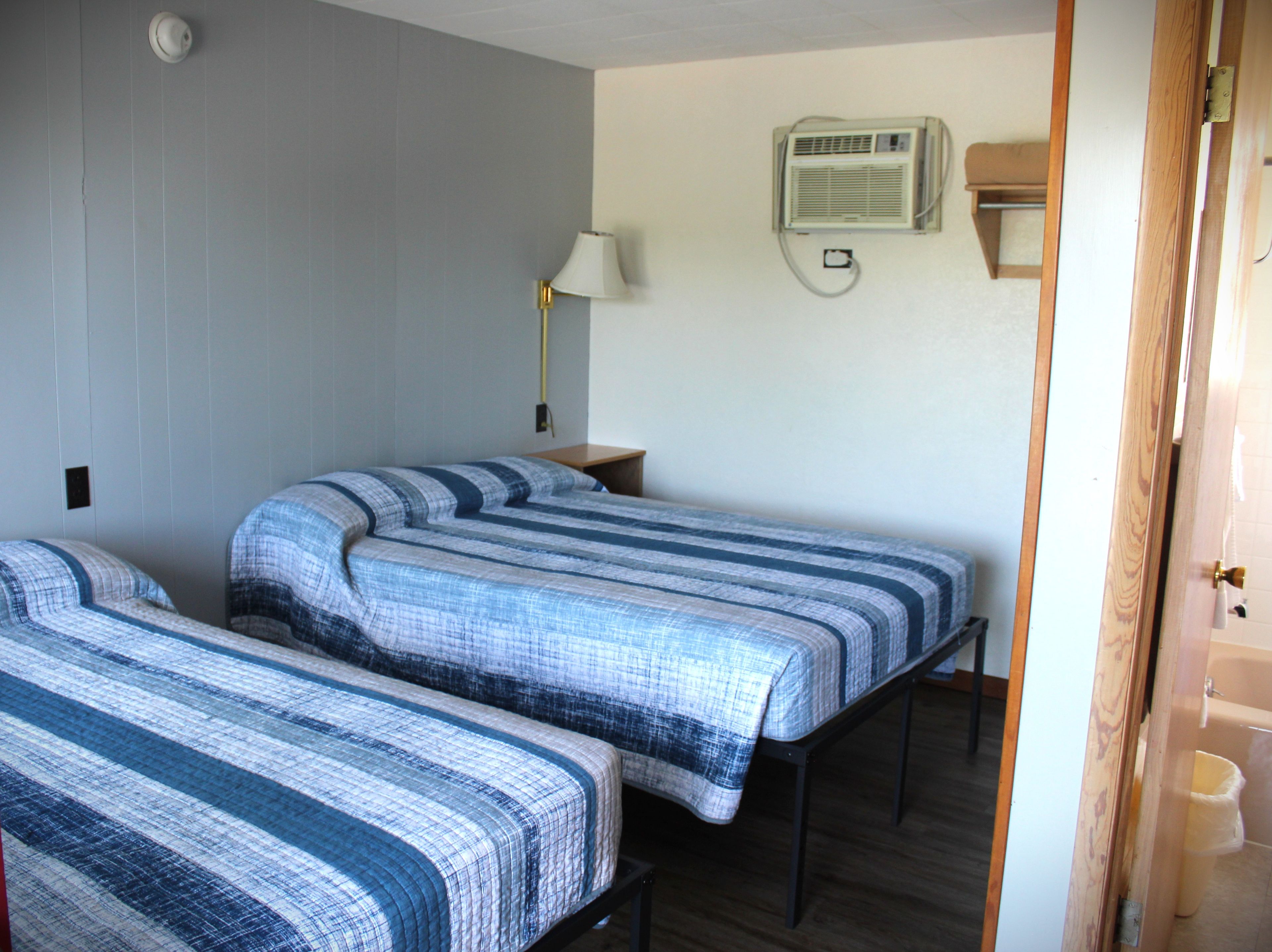A room with two beds, a nightstand, a lamp, and a wall-mounted air conditioner.