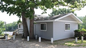 A small, single-story cabin with a sloped roof, a front porch, and a tree in front.