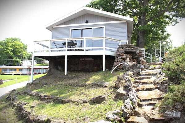 A two-story house with a balcony, a stone staircase, and a view of a body of water in the background.