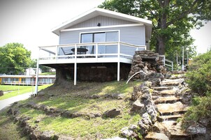 Una casa de dos pisos con un balcón, una escalera de piedra y vista a un cuerpo de agua al fondo.