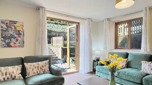 Sala de estar com sofá, mesa de centro e um vaso de flores. Há um quadro na parede, uma janela com cortinas e uma porta que dá para o exterior.