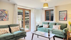 Uma sala de estar com dois sofás verdes, uma mesa de centro e vista para um jardim através de uma porta de vidro.