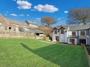 A stone building with a wooden fence, a grassy yard, and a white house with a wooden door. - Ashton Cottage, Countisbury (Lynmouth)