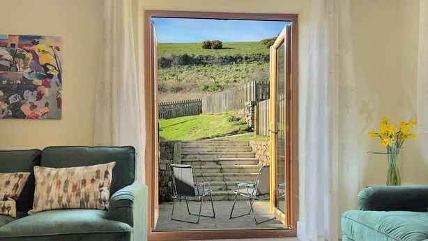 Quarto com sofá verde, uma pequena mesa e vista para uma área verde externa através de uma porta.