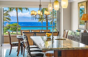 A modern kitchen with a large island, dining area with chairs, and a view of the ocean. - Oceanfront 3BR Corner Suite with Wraparound Lanai and Private BBQ Grill (Lahaina)
