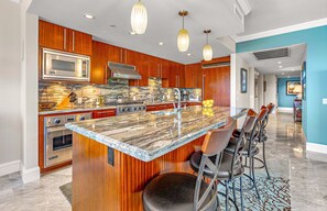 A modern kitchen with a marble island, wooden cabinets, stainless steel appliances, and a bar area with stools. - Oceanfront 3BR Corner Suite with Wraparound Lanai and Private BBQ Grill (Lahaina)