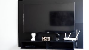 Un téléviseur à écran plat fixé sur un mur noir, avec un routeur blanc et un petit objet blanc posé sur une étagère noire en dessous.