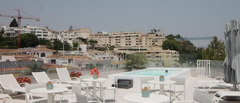 A rooftop terrace with lounge chairs and tables, a swimming pool, and a view of a cityscape with buildings and a bridge.