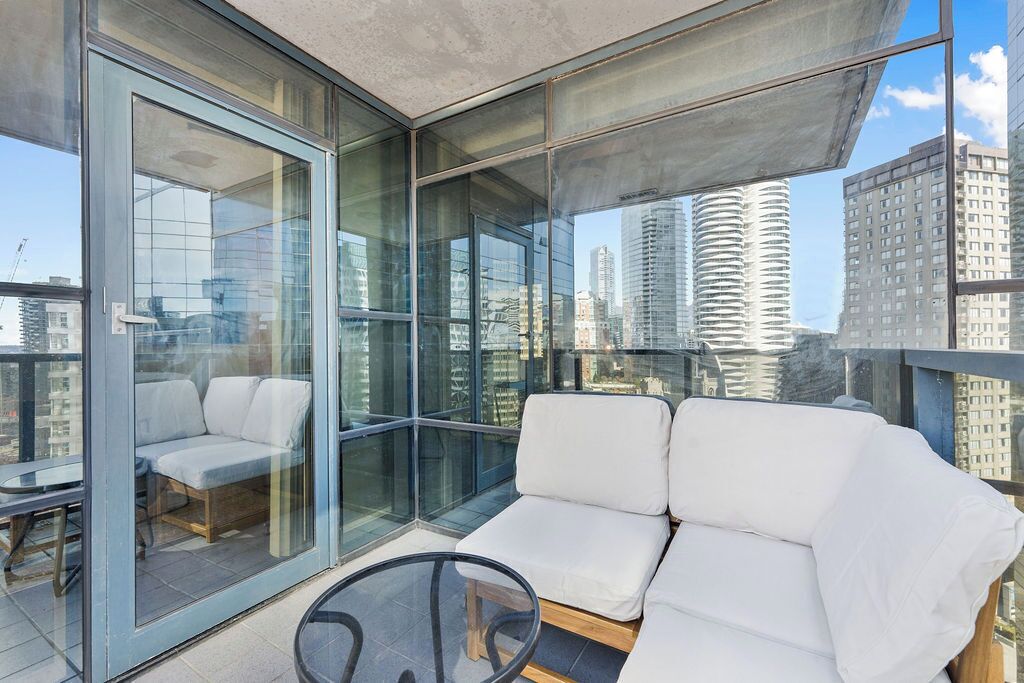 A modern balcony with glass walls, a cushioned seating area, and a view of high-rise buildings.
