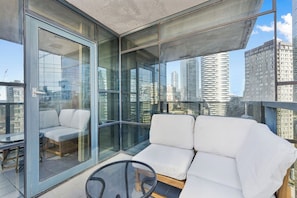 Un balcon moderne avec des murs en verre, un coin salon confortable et une vue sur les gratte-ciel.