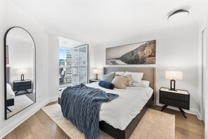 Une chambre moderne avec un grand lit, une vue sur la ville, un grand miroir et un tableau accroché au mur.