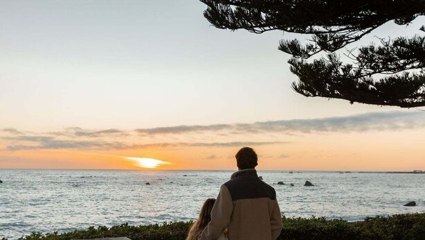 A couple sitting at a wooden table overlooking the ocean during sunset. - Beach Nearby | Balcony | Stroll to Town – KK77018 (Kaikōura)