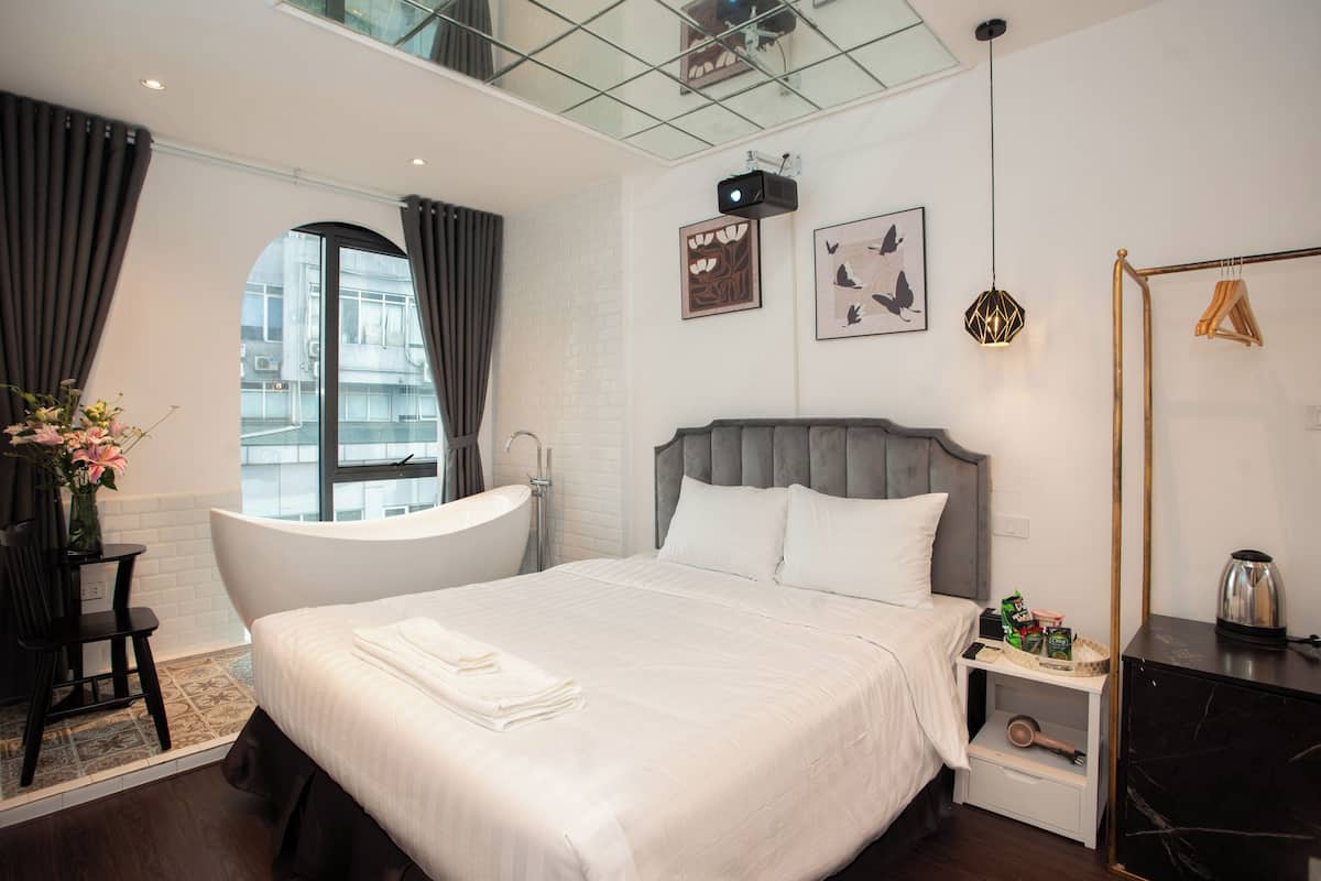 Een moderne hotelkamer met een groot bed, een badkuip, een klein tafeltje met toiletartikelen en uitzicht op de stad door een raam.