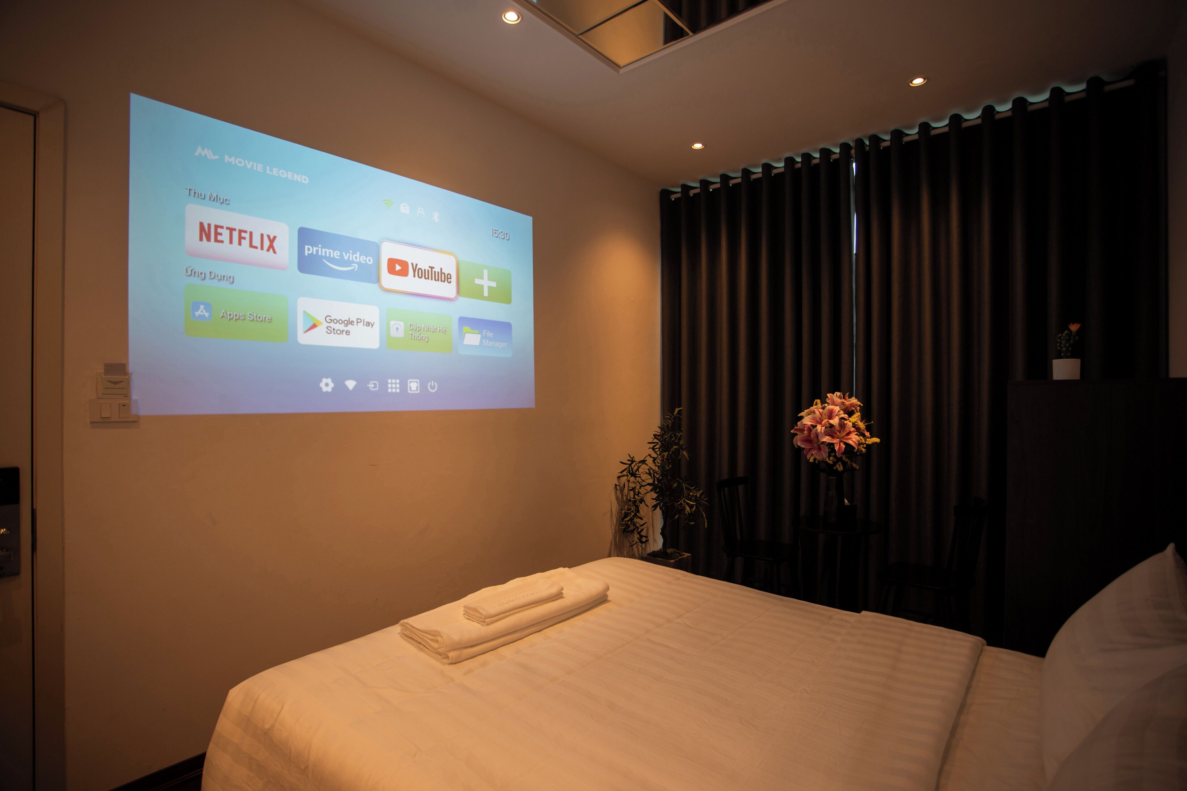 Une chambre d’hôtel avec un lit, un écran de projection diffusant des services de streaming, une plante en pot et un rideau foncé.
