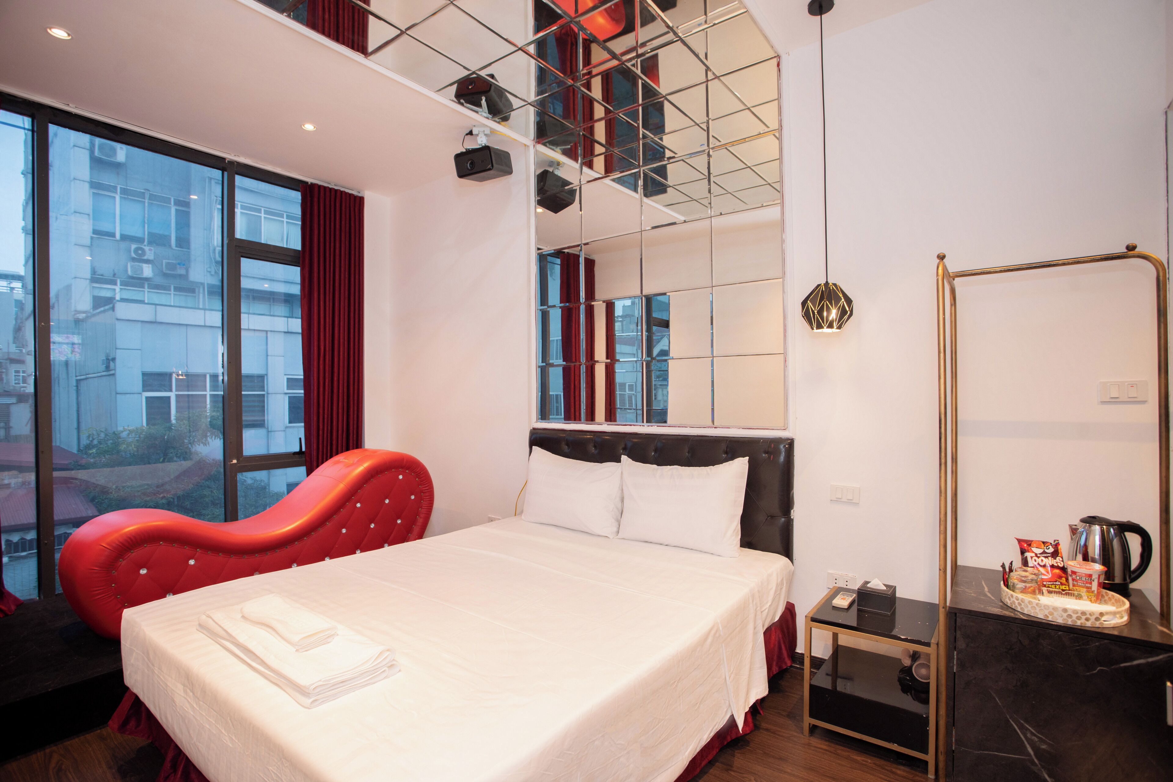 Habitación de hotel con cama, una silla roja, una mesita con una tetera y vistas a edificios a través de amplias ventanas.