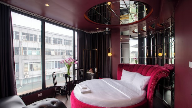 A hotel room with a large bed, a round seating area, a desk, and a view of a building through large windows. - Luma Boutique Hotel (Hanoi)