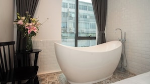 A modern bathroom with a freestanding bathtub, a window with curtains, and a vase with flowers on a table. - Luma Boutique Hotel (Hanoi)