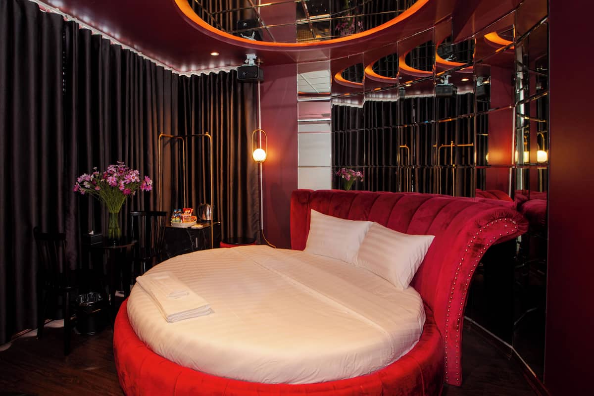 Een rond bed met een rood beklede hoofdsteun, een nachtkastje met een lamp en een spiegel aan de muur.