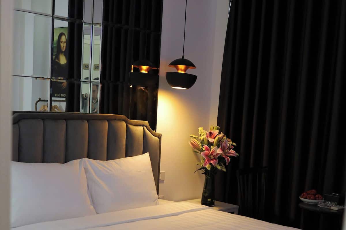 Een keurig opgemaakt bed met witte lakens, een modern hoofdbord, een nachtkastje met een bloevas en een spiegel die een deel van de kamer weerspiegelt.