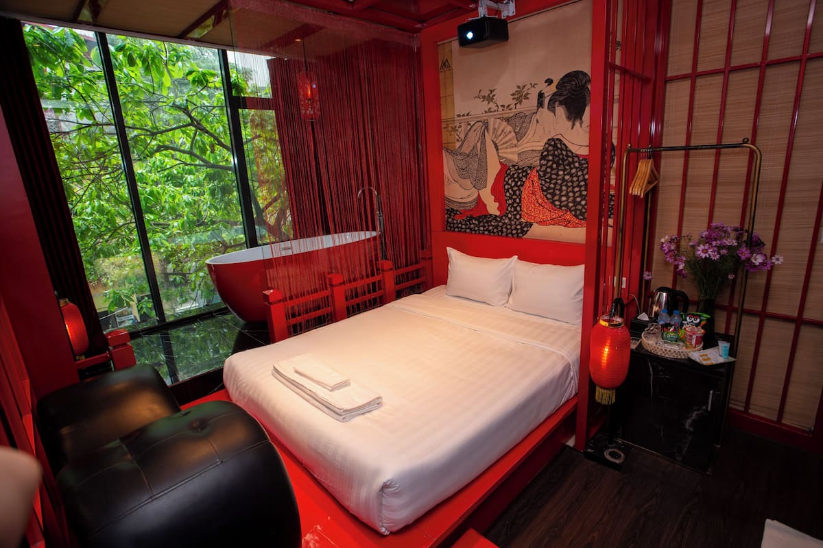 Een moderne hotelkamer met een groot bed, een rode stoel, een tafeltje met een bloevas en een groot raam met uitzicht op groen.