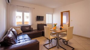Un salon avec un canapé en cuir marron, une table à manger avec des chaises, un téléviseur à écran plat fixé au mur et une fenêtre avec des rideaux.