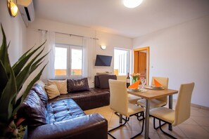 Ein Wohnzimmer mit einer Ledersofa, einem Esstisch mit Stühlen, einem an der Wand befestigten Flachbildfernseher und einer großen Pflanze.