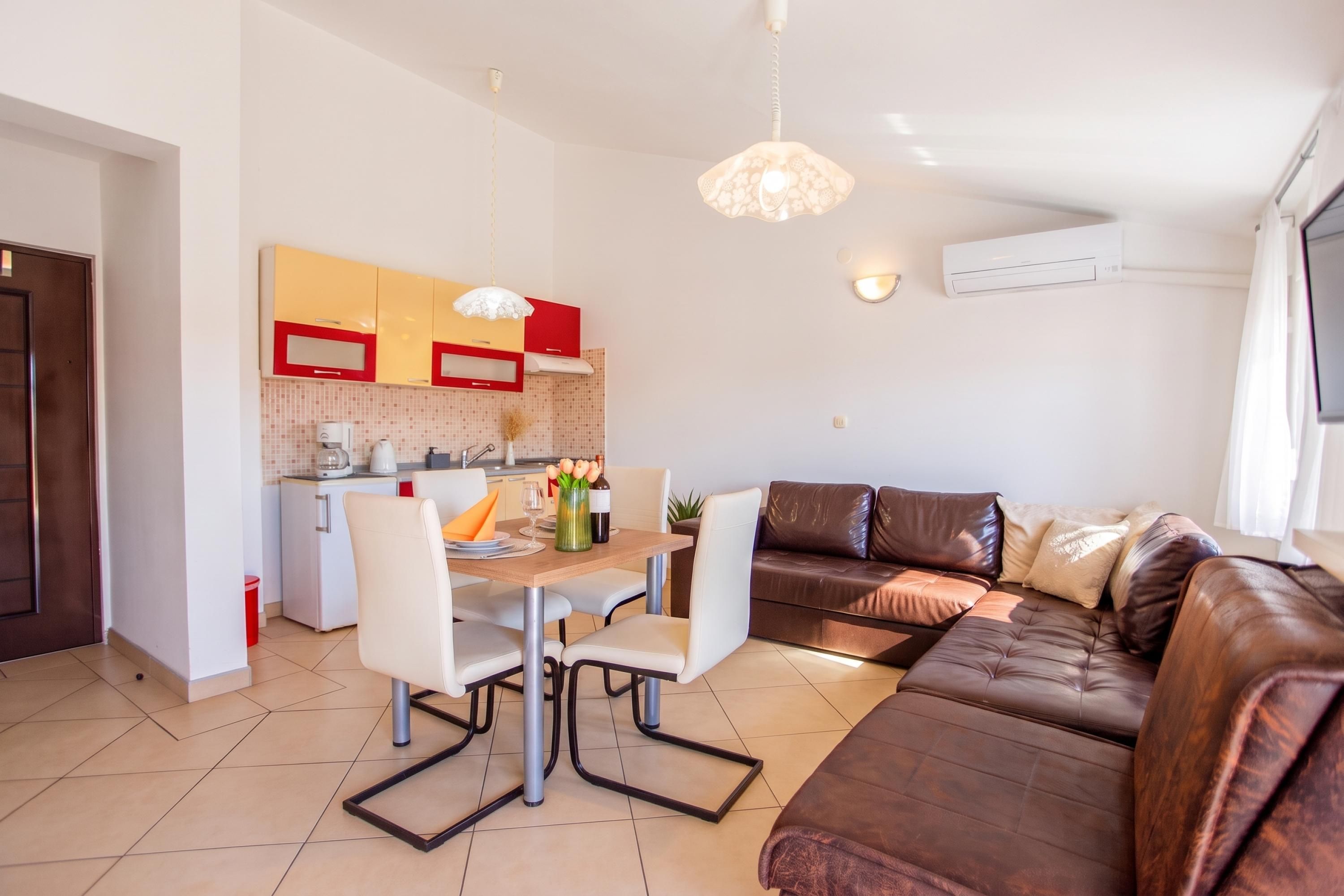 A modern living room with a brown leather sofa, a dining table with chairs, a kitchen area with red and yellow cabinets, and a pendant light.