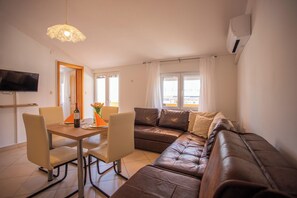 Un salon comprenant un canapé, une table à manger avec des chaises, un téléviseur fixé au mur et une fenêtre avec des rideaux.