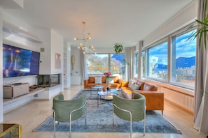 A modern living room with a large TV, comfortable seating, a coffee table, and a view through large windows.