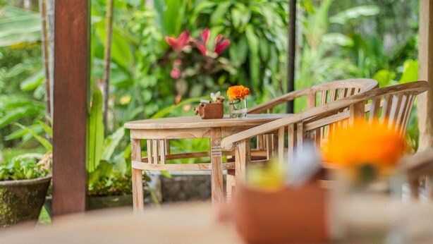 A wooden table and chair set outdoors with a glass of orange juice and a vase with flowers. - D'junas Villas (Ubud)