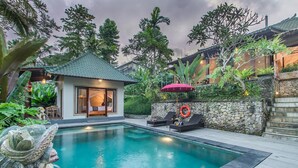 A pool area with a small house, lounge chairs, and a stone wall. - D'junas Villas (Ubud)