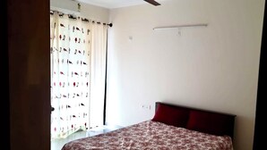 A bedroom with a bed, a ceiling fan, a window with curtains, and a wall-mounted light. - Enjoyable 3-bedroom apartment with AC in vibrant Bengaluru (Bengaluru)