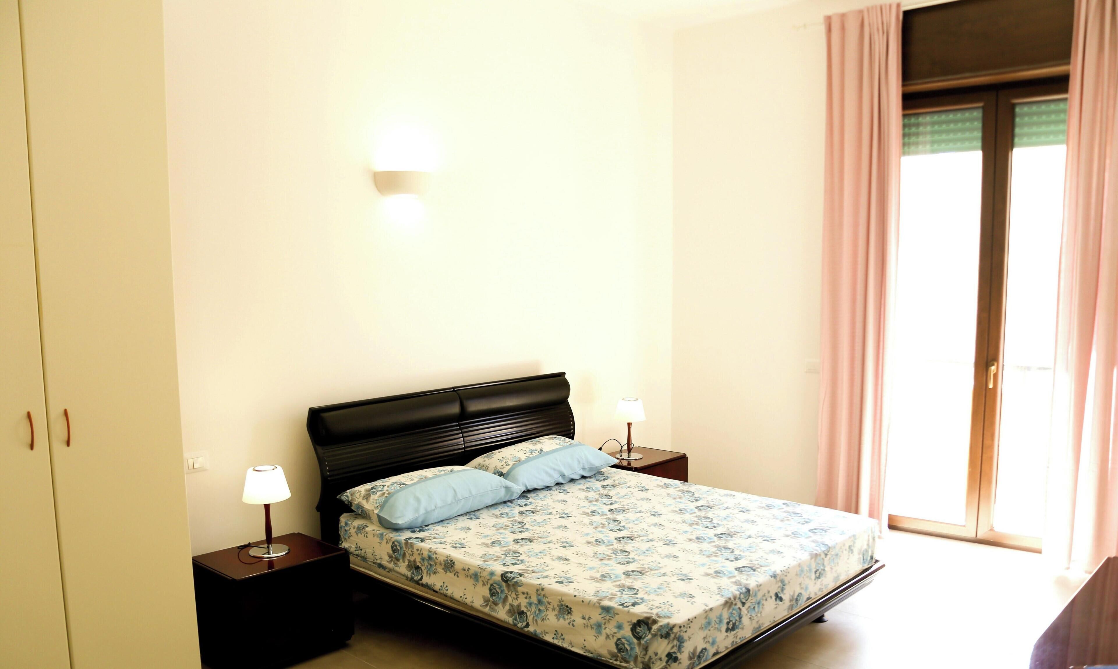 Ein Schlafzimmer mit einem Bett, zwei Nachttischen, einer Wandleuchte und einem Fenster mit Vorhängen.