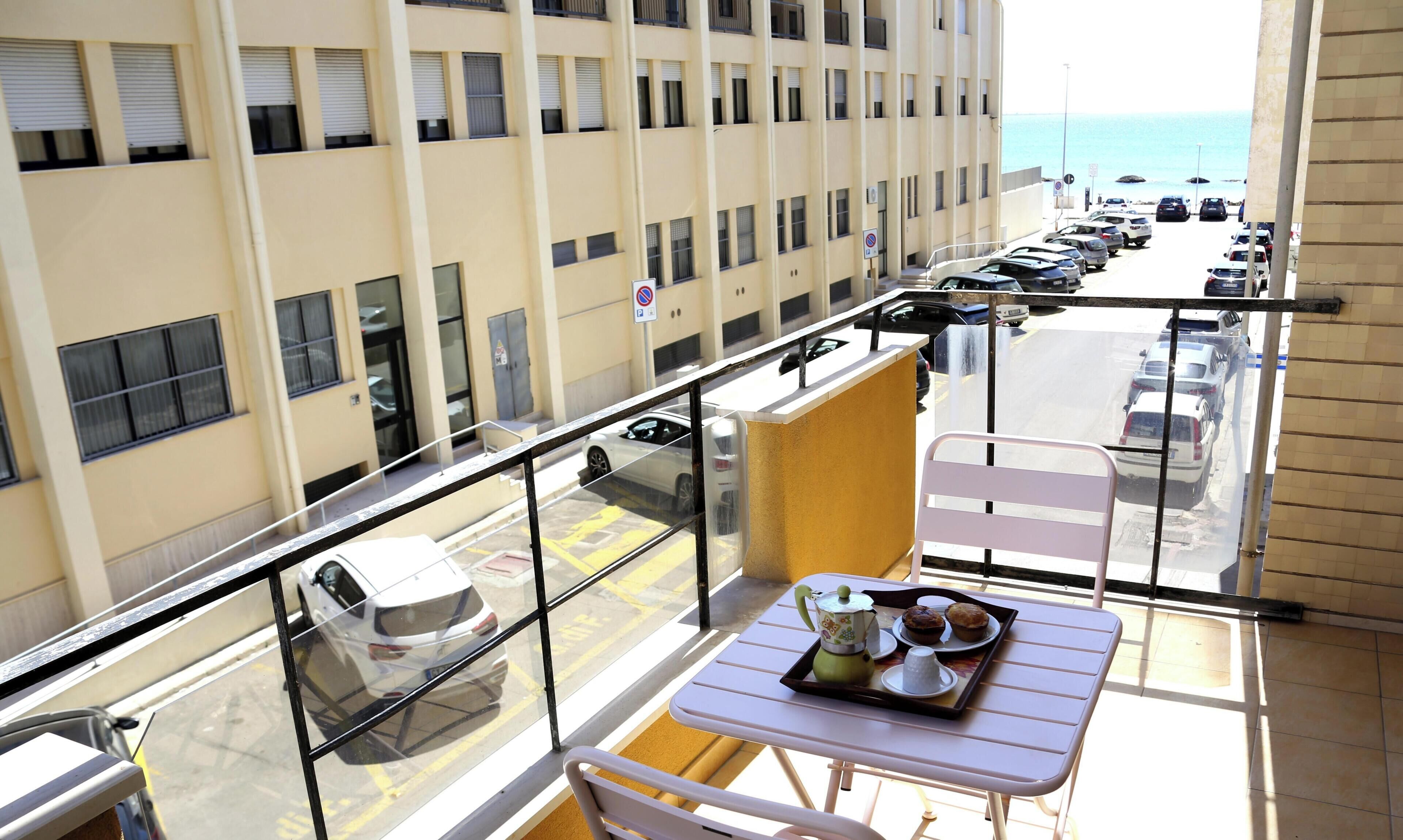 A balcony with a table set for tea, a view of parked cars, and a building with multiple windows.