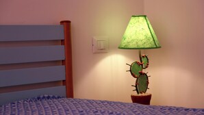Una cama con una colcha azul estampada, un cabecero de madera, una lámpara de cactus verde y un interruptor blanco en la pared.